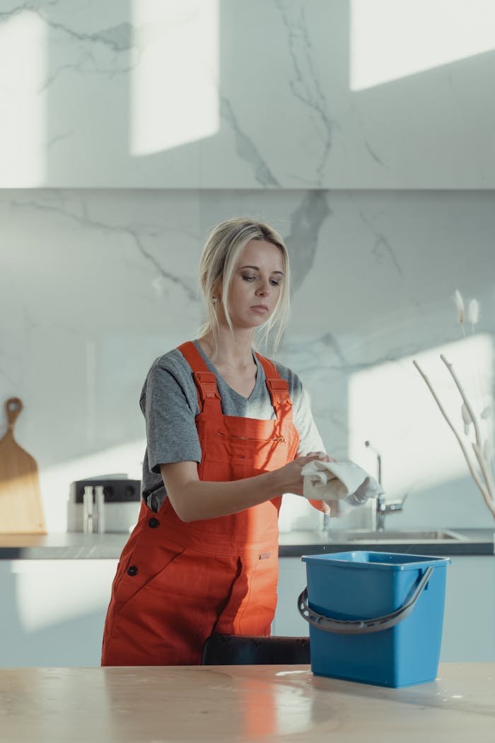 portfolio-03 A woman wearing orange overalls cleans a kitchen counter with a rag and pail in a modern kitchen.
