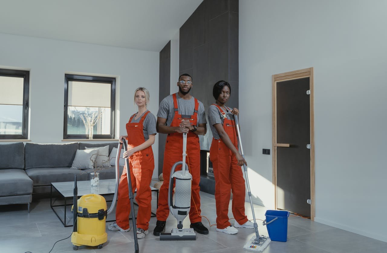 portfolio-04 Diverse cleaning team in red uniforms ready to sanitize a modern living room with vacuum and mop.